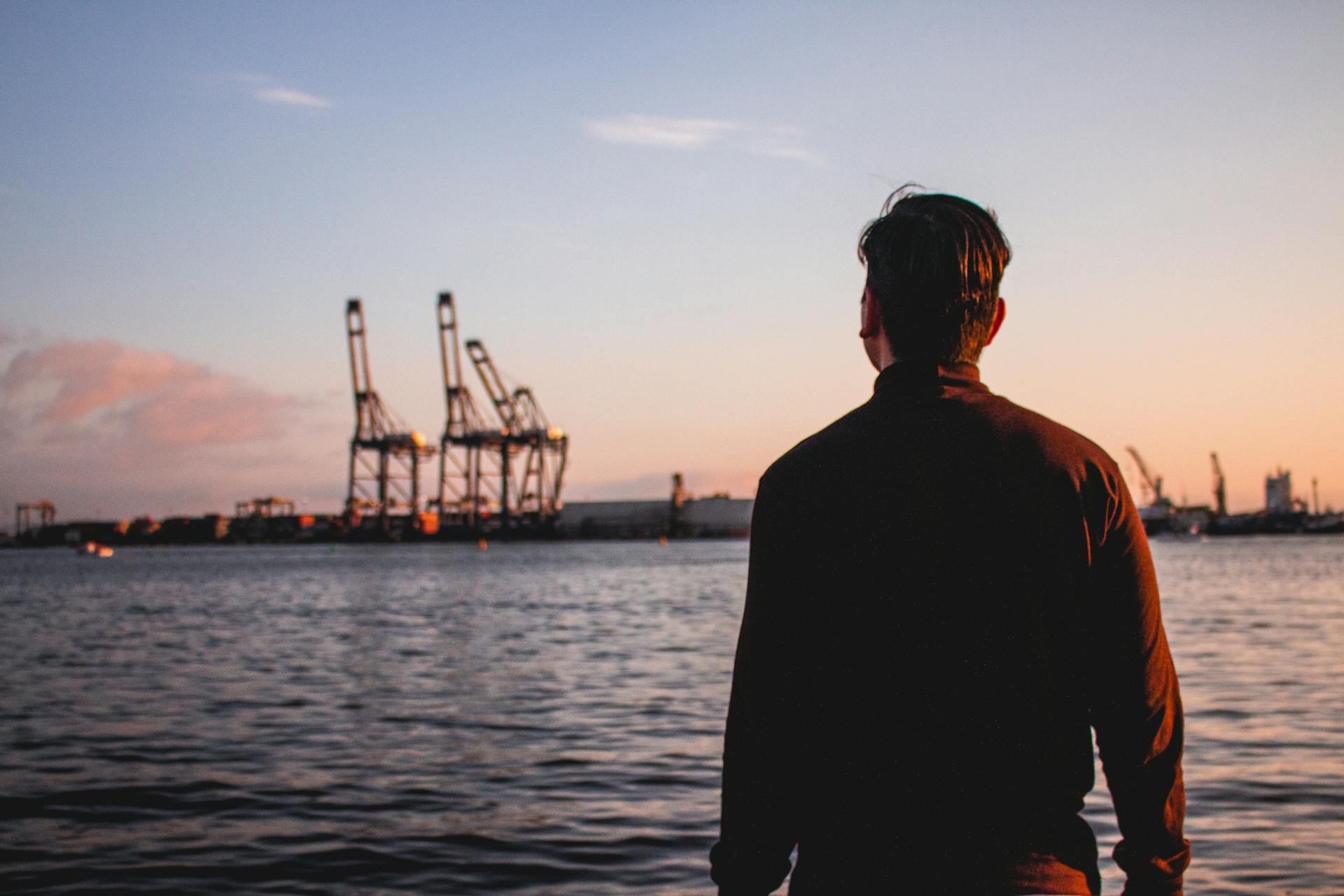 A man stands at a port watching the sunset over the water with industrial cranes in the distance.