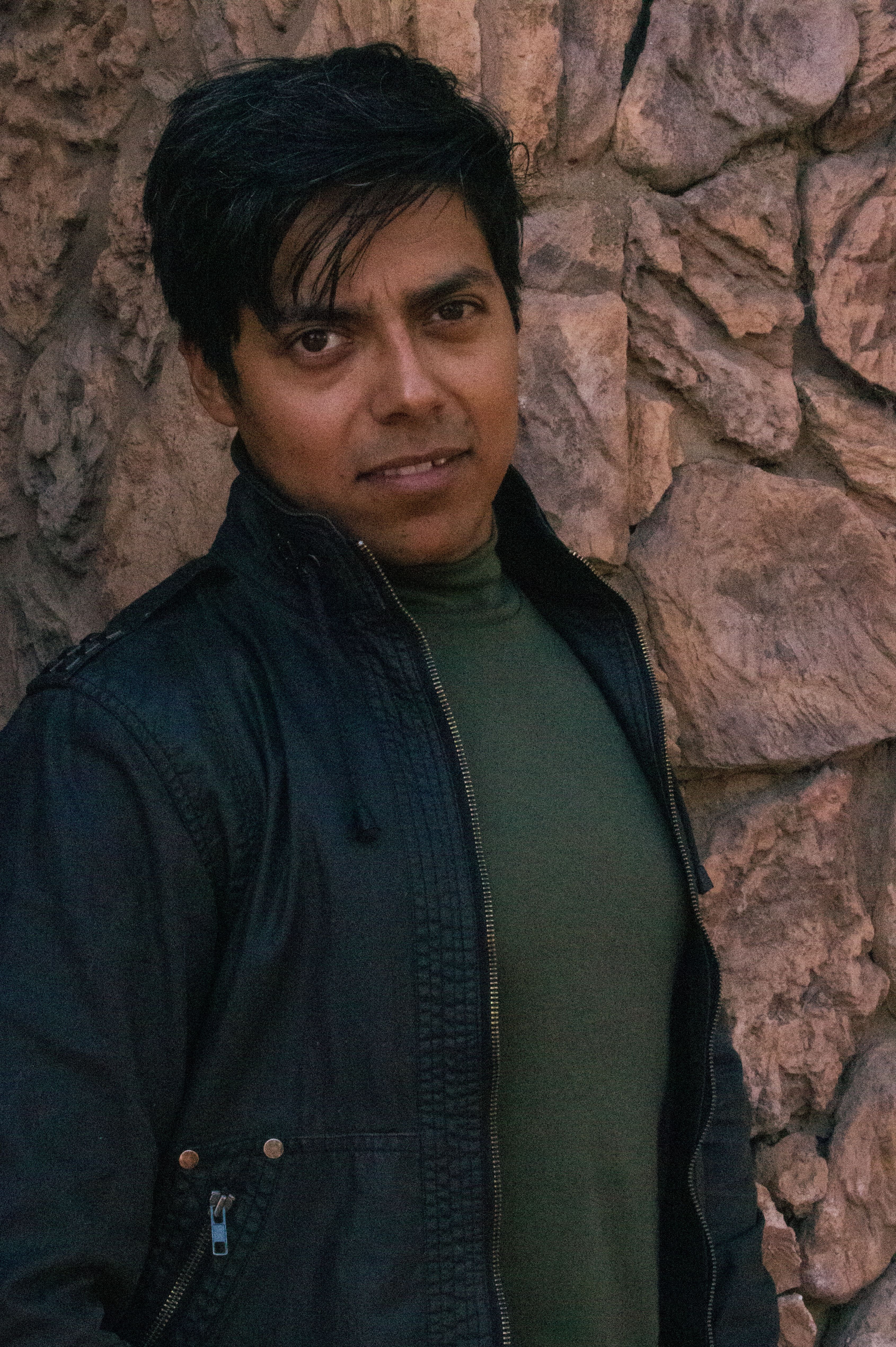 Stylish portrait of a man in a black jacket posing against a textured stone wall.