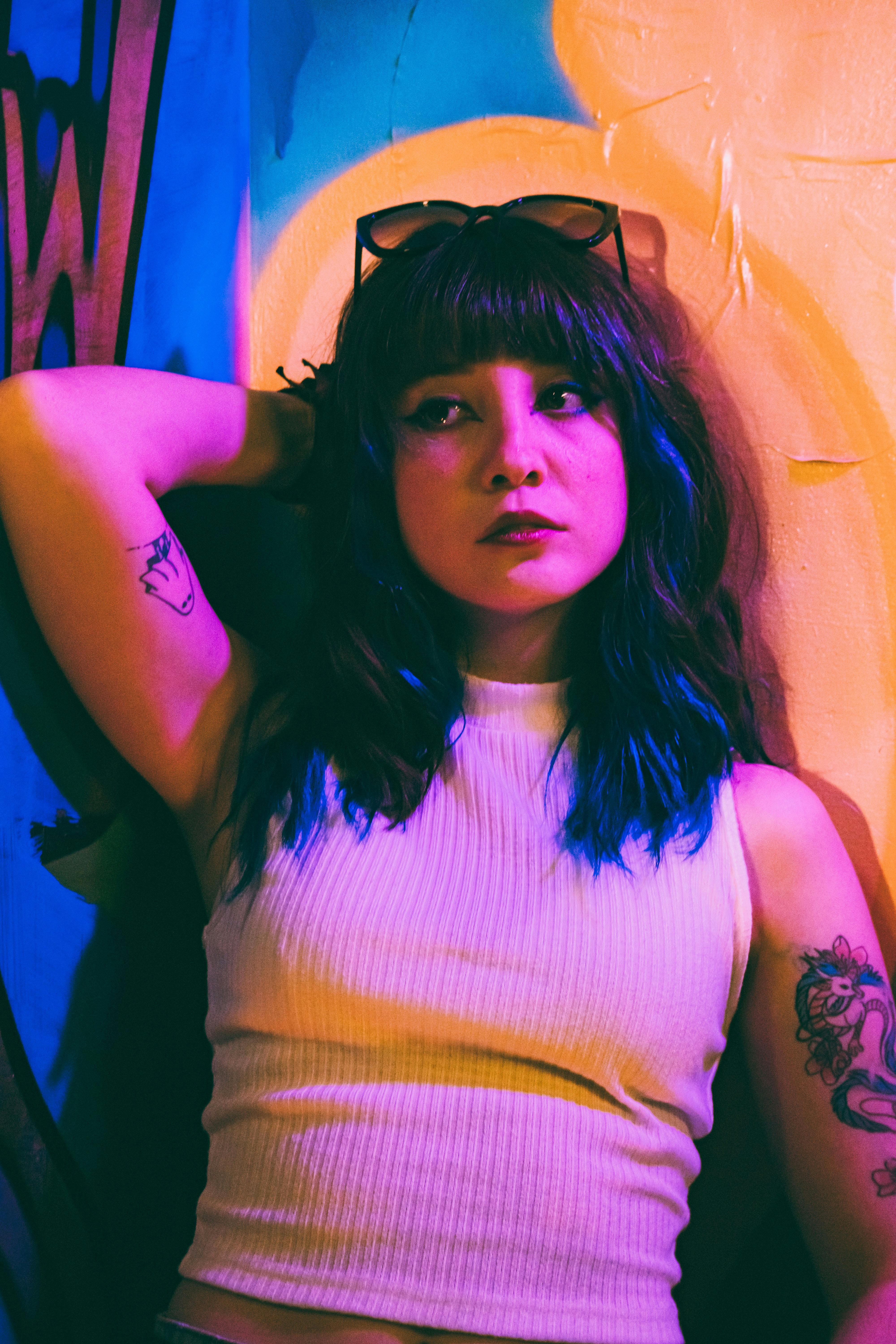 Stylish woman with tattoos poses against vibrant urban backdrop, illuminated by colorful lighting.
