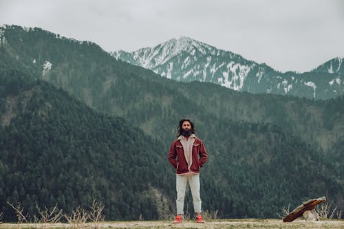 A Man in Red Jacket Standing Near the Mountain with Green Trees