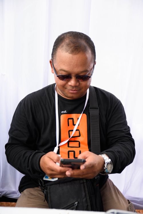 Free Asian man wearing a black sweater and sunglasses sits indoors while using a smartphone. Stock Photo