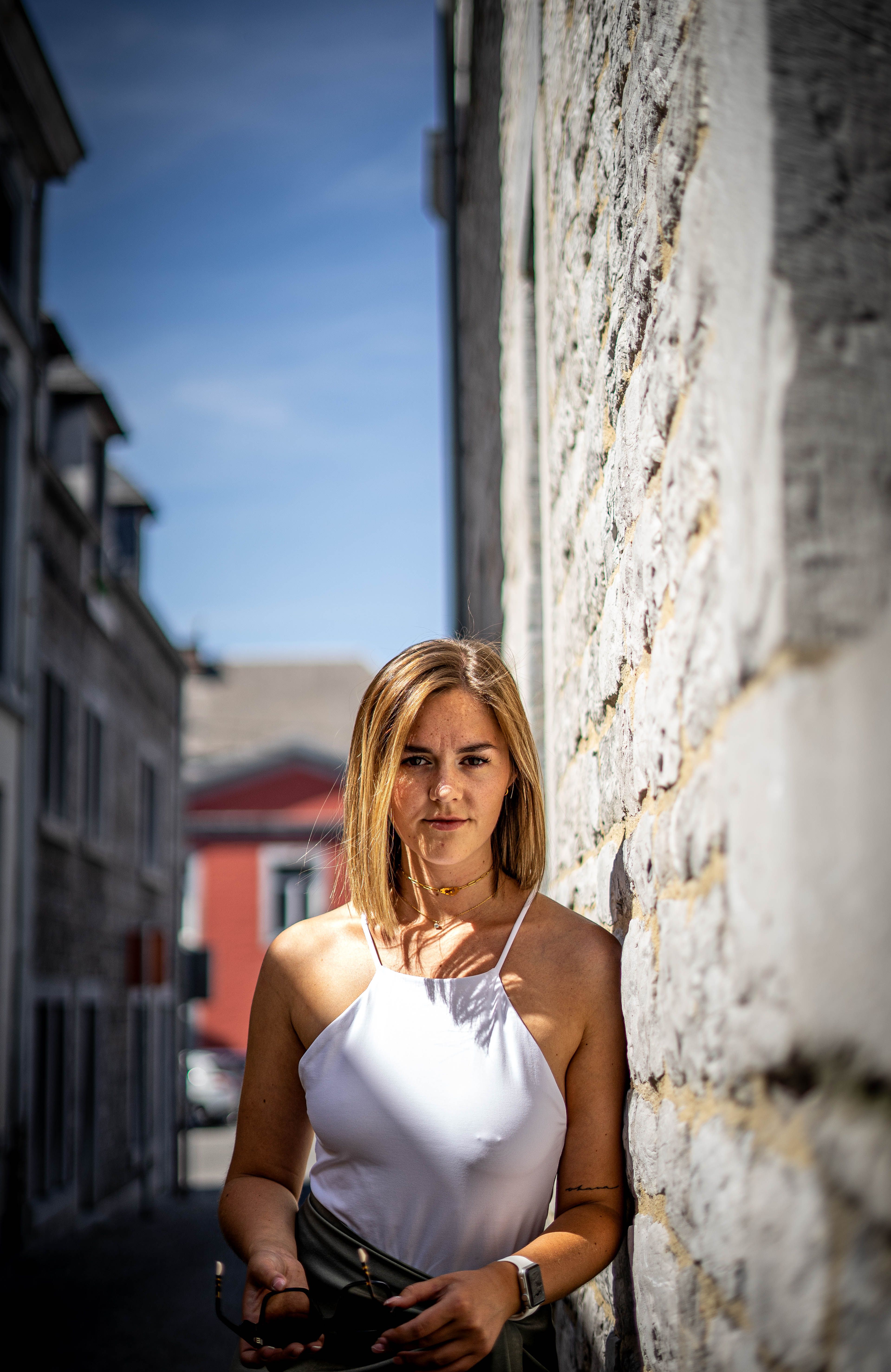 A young woman with blonde hair in a white tank top leaning against a stone wall in the street.