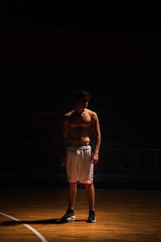 Muscular shirtless man standing on a dimly lit basketball court, emphasizing fitness and sports theme.