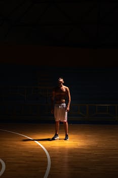 A shirtless man stands in a spotlight on a basketball court.