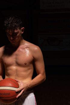 A shirtless male athlete holding a basketball under moody lighting with a black background.