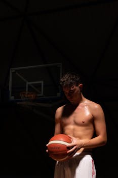 A shirtless young man holding a basketball in a dimly lit indoor court.