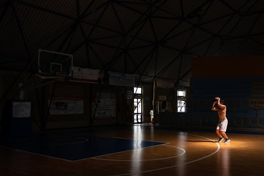 Shirtless athlete practicing basketball shots alone in a dimly lit gym.