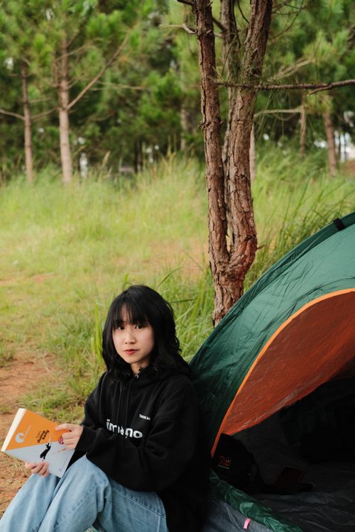 Free Asian woman reading while camping, sitting by a tent amidst nature. Stock Photo