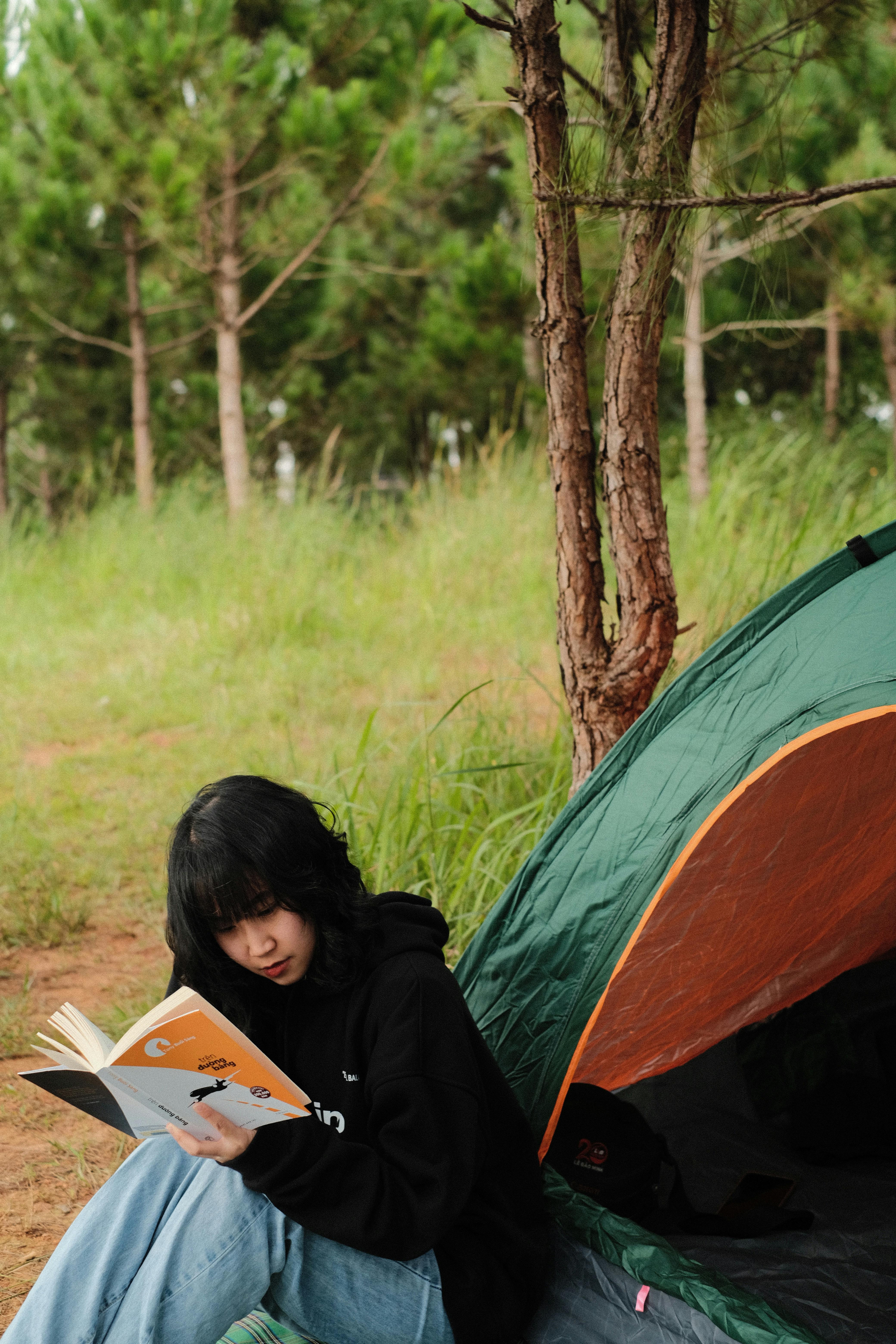 Woman enjoys a peaceful moment reading next to a camping tent surrounded by pine trees.