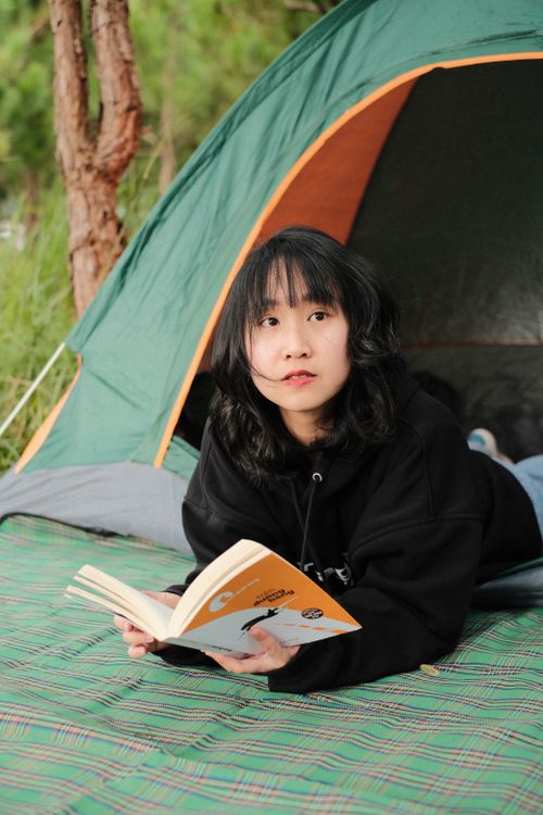 Free Young woman enjoys reading a book while relaxing outside a camping tent in a natural setting. Stock Photo