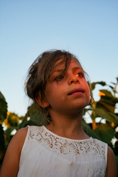 A young girl gazes thoughtfully in a sunlit garden, wearing a white dress.