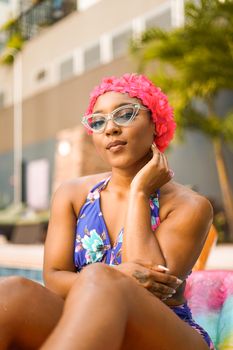 Trendy woman in floral swimsuit and pink swim cap lounging by the poolside on a sunny day.