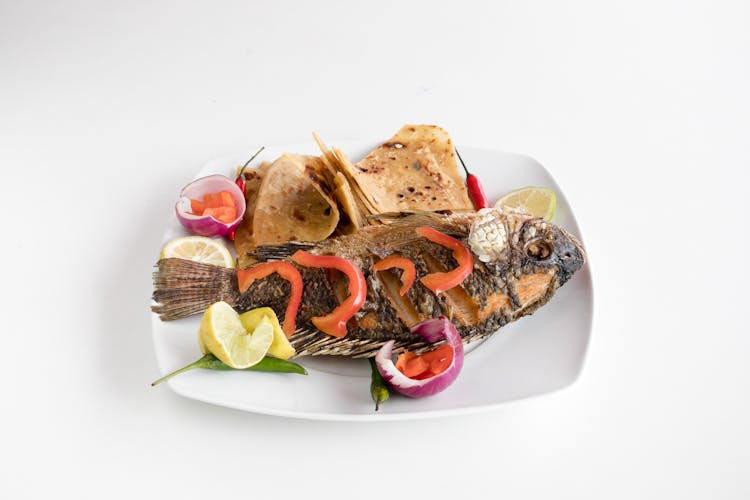 A Fried Tilapia On Plate With Tomatoes, Onions And A Slice Of Lemon