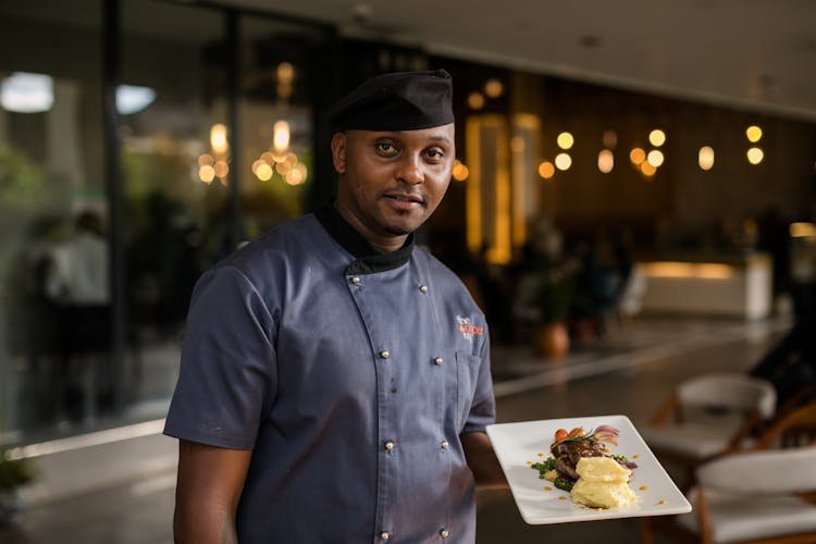 A Man In Blue Chef's Uniform Holding A Plate Of Delicious Food