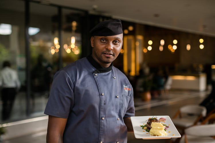 A Man In Blue Chef's Uniform Holding A Plate Of Delicious Food
