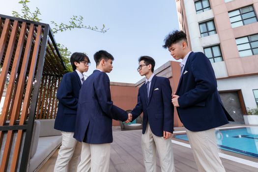Four teenage boys in school uniforms shaking hands on a rooftop terrace.