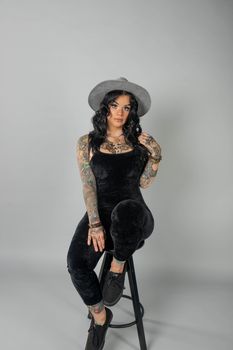 Portrait of a tattooed woman sitting on a stool wearing a hat, showcasing unique fashion sense.