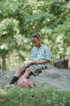 A young boy enjoys reading a book on a rock in a park, surrounded by greenery.