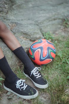 A colorful football on grass beside a person's legs in casual attire, outdoors.