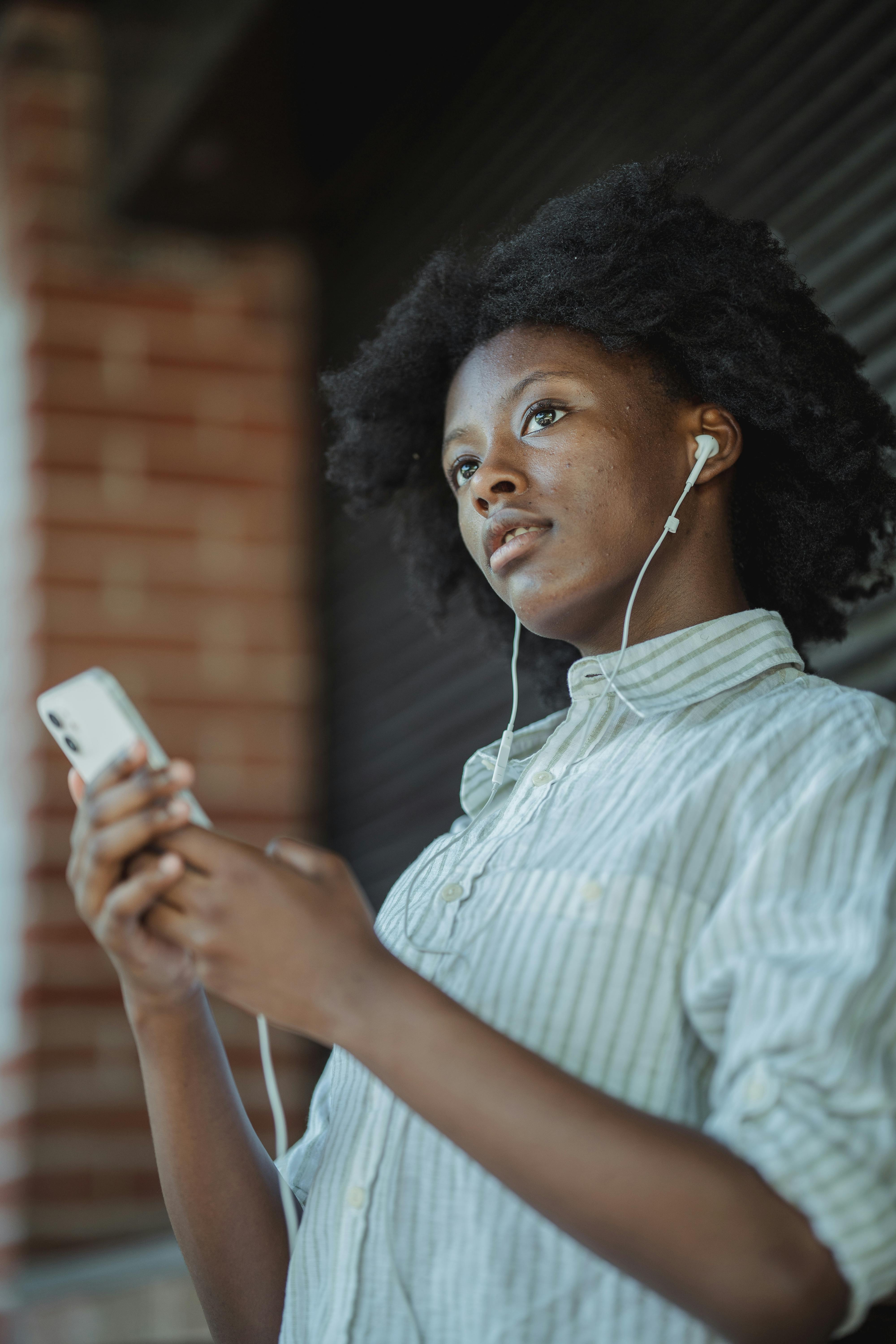 Free A young woman enjoys music on her smartphone using earphones, leaning against a brick wall. Stock Photo
