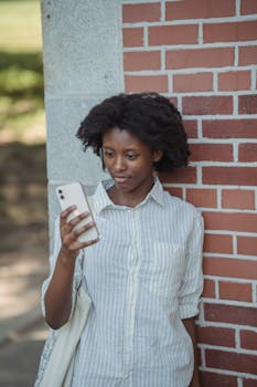 A young woman stands against a brick wall, engrossed in her smartphone outdoors.