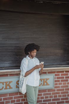 A young woman stands outdoors, listening to music on her smartphone, leaning against a brick wall.
