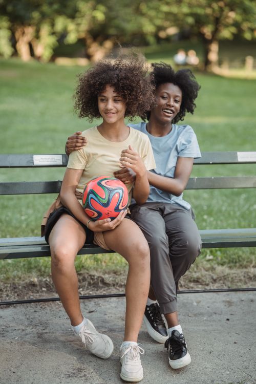 Free Two teenagers enjoy a sunny day outdoors, sitting on a bench and embracing while holding a soccer ball. Stock Photo