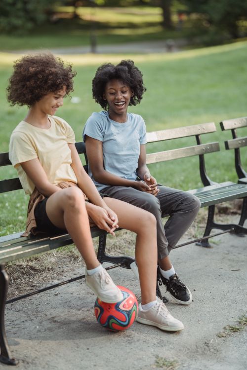 Free Two girls sitting on a park bench smiling and playing with a soccer ball outdoors. Stock Photo