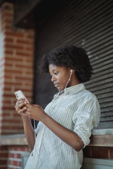 A woman leaning against a building, listening to music on her smartphone with earphones.