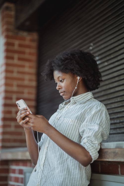 Free A woman leaning against a building, listening to music on her smartphone with earphones. Stock Photo