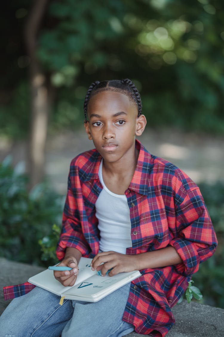 Portrait Of A Boy With Braided Hair And Red Checked Shirt Sitting In A Park And Drawing In A Notebook