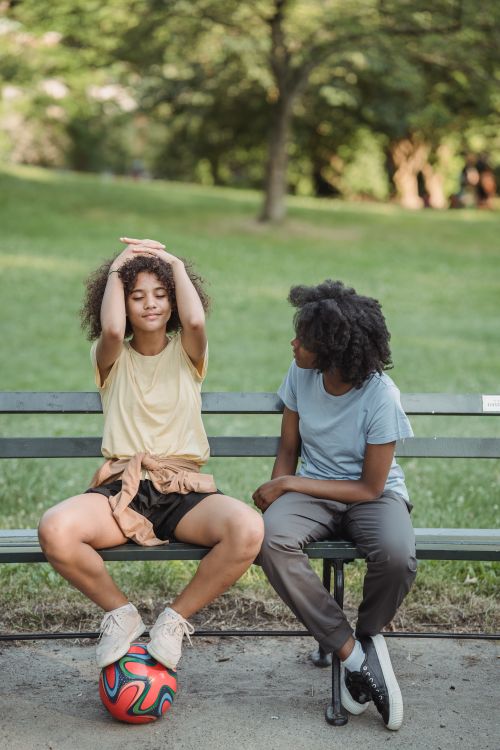 Free Two teenagers sitting on a park bench with a ball, enjoying a sunny day outdoors. Stock Photo