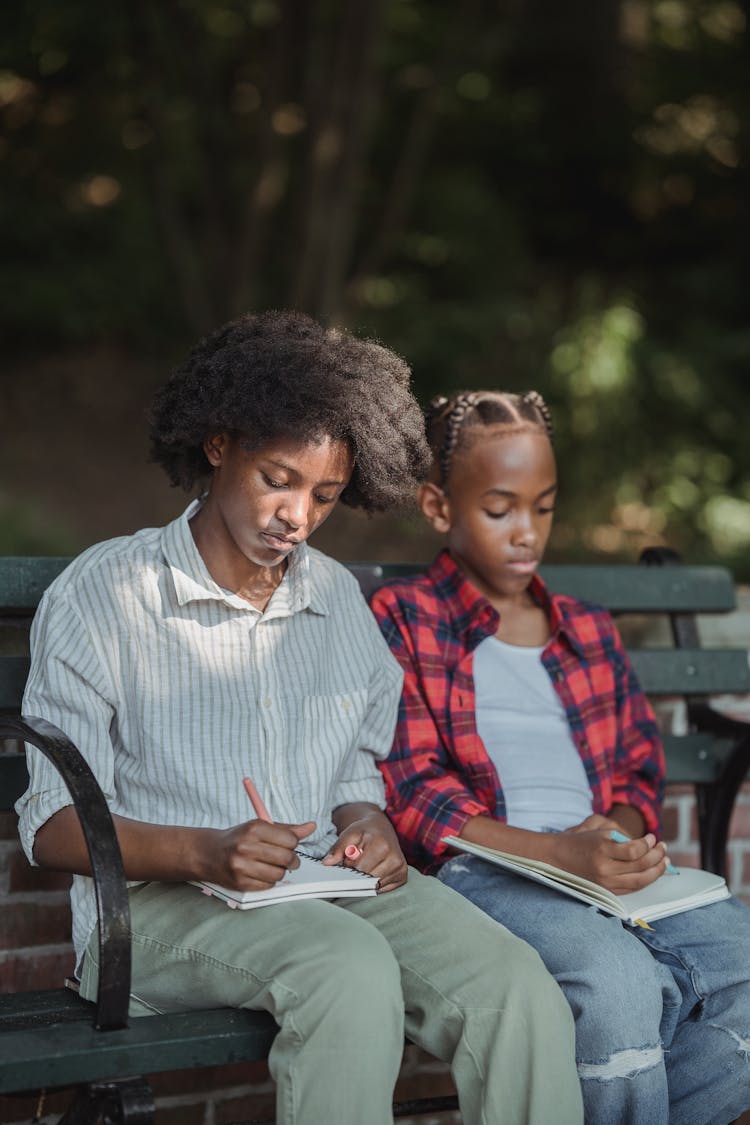 Girl And Boy Sitting On A Bench In A Park And Drawing In Notebooks