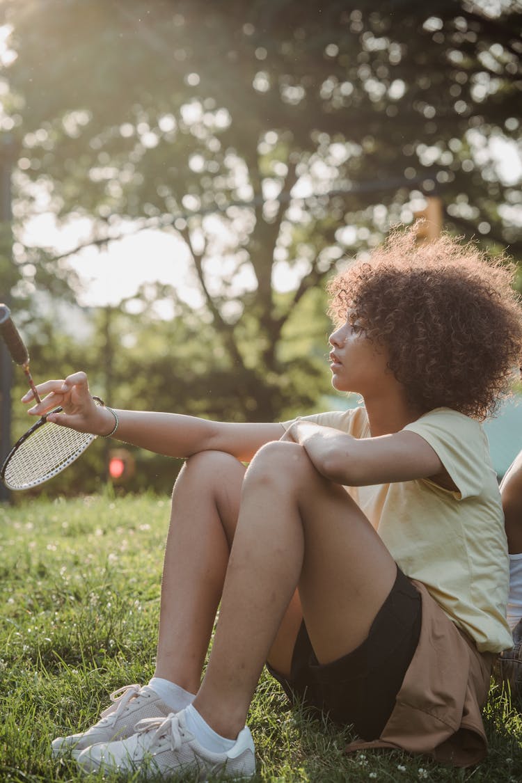 Profile Of A Girl With Brown Curly Hair Sitting In A Park With A Badminton Rocket