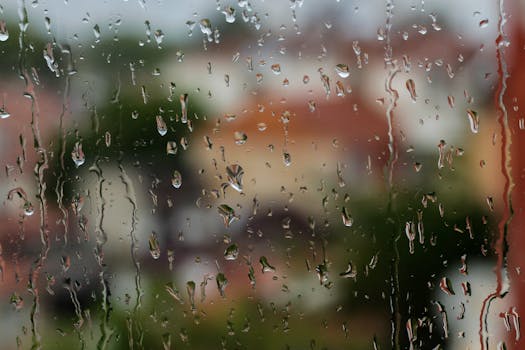 Rain droplets on a window with a blurred view of houses and greenery outside.