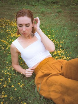 A woman with red hair relaxing on grass with yellow flowers, wearing a white camisole and orange pants.