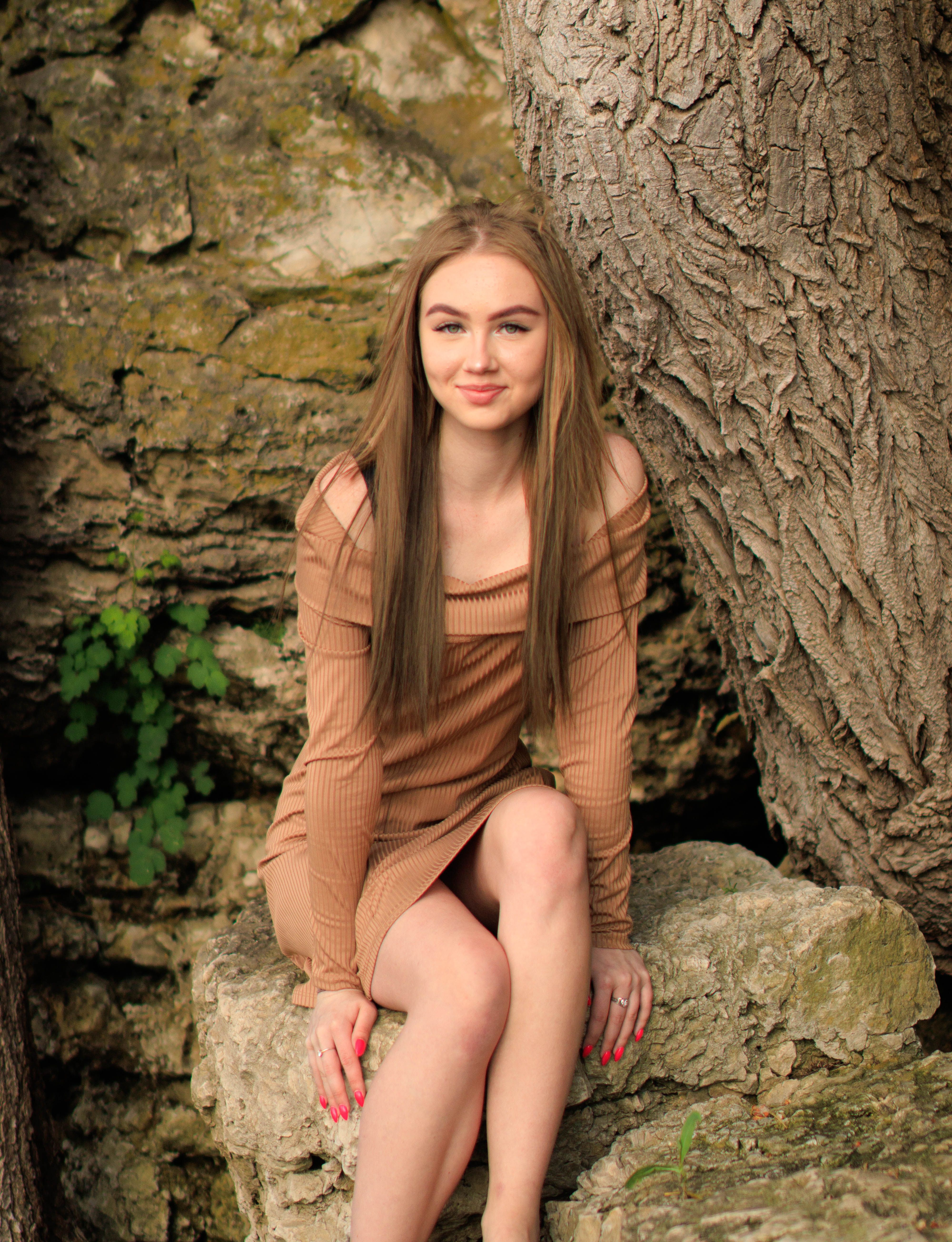 A young woman in a brown off-shoulder dress sits on a rock with a natural backdrop.