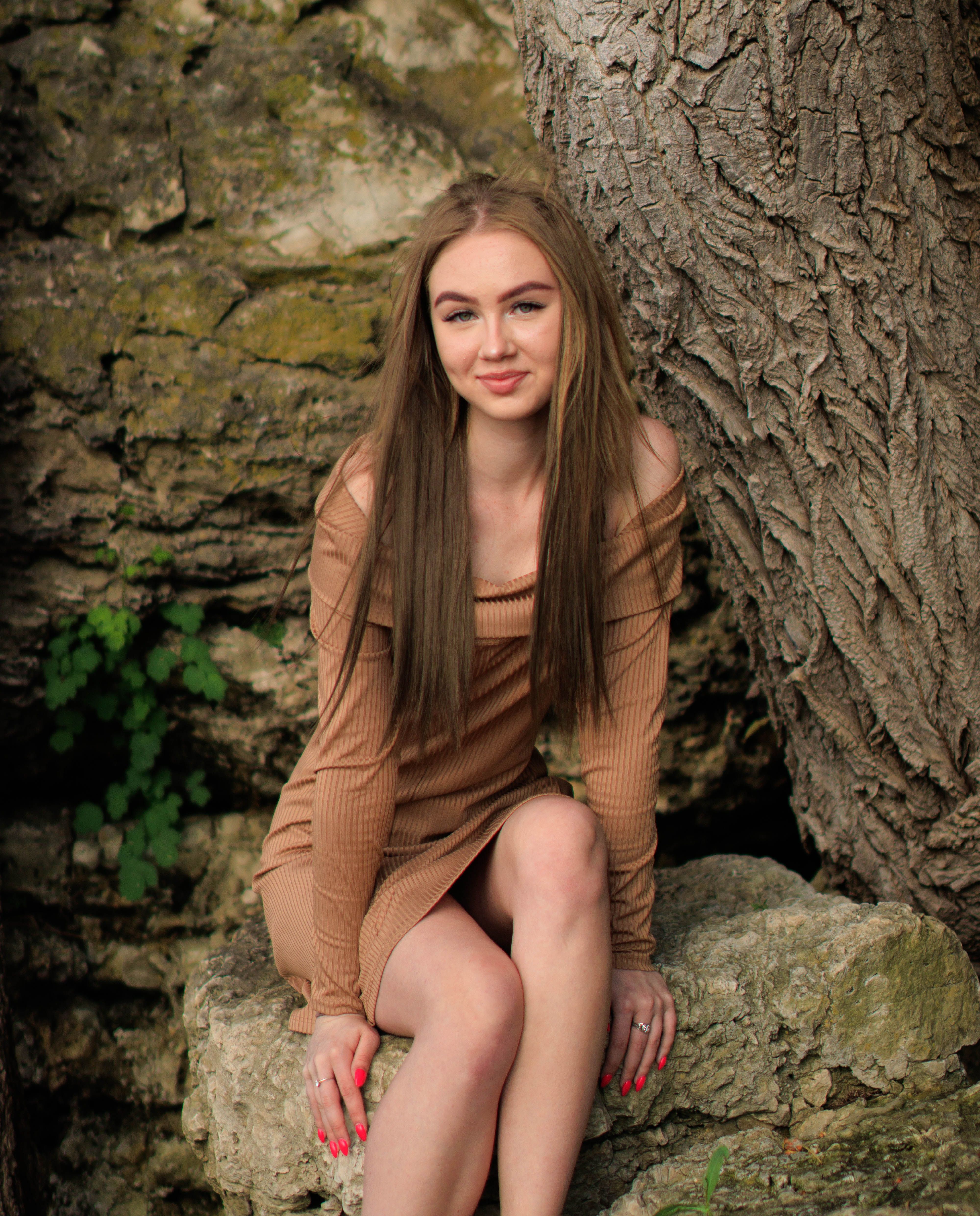 A young woman in an off-shoulder brown dress sitting on a rock, smiling.