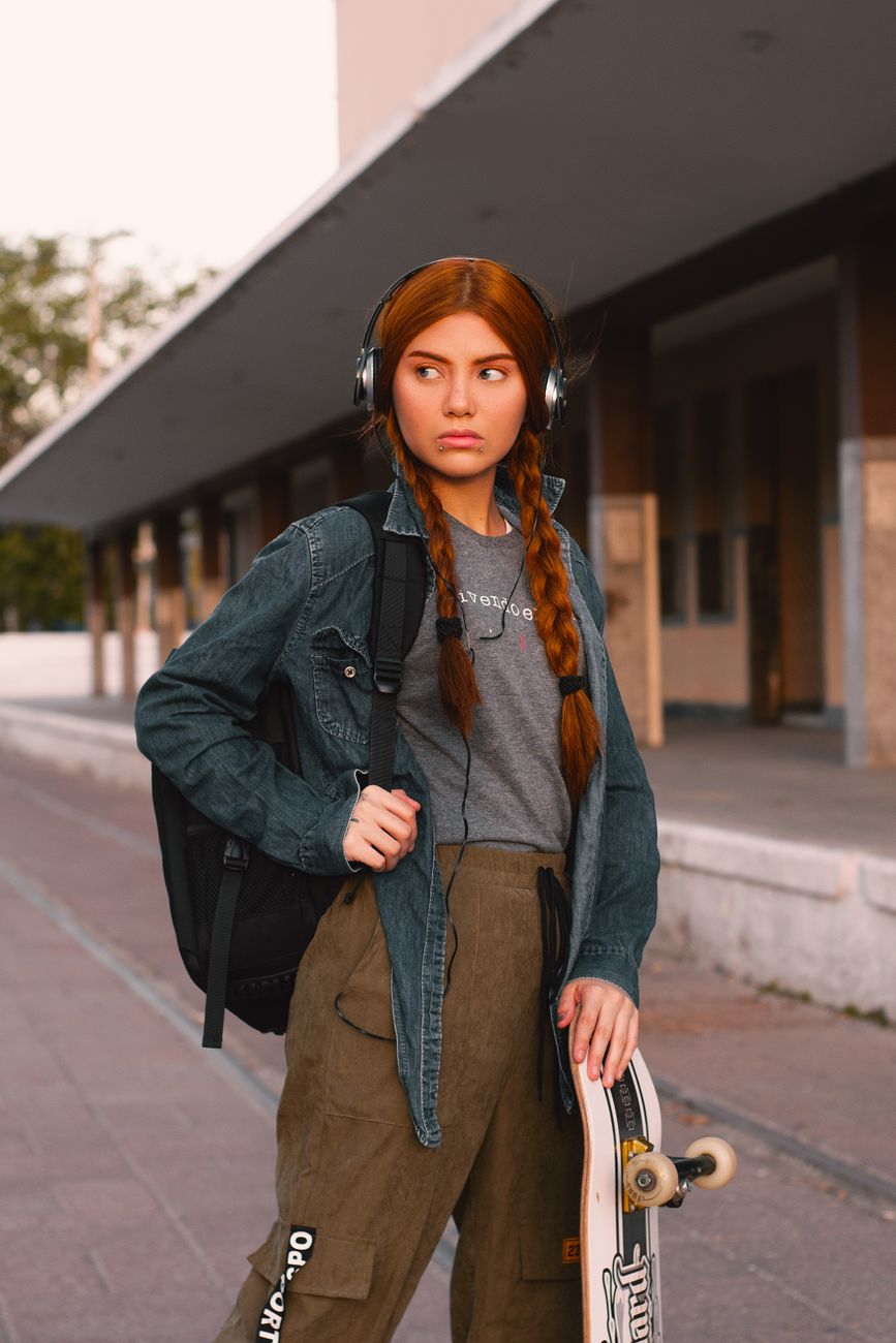 Stylish redhead woman in urban setting, wearing a denim jacket and holding a skateboard.