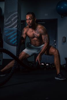 A muscular tattooed man intensely working out with battle ropes indoors.