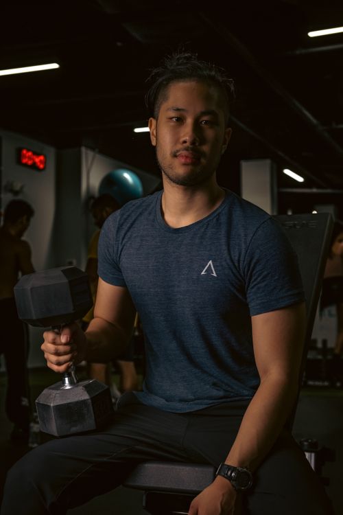 Free Asian man sitting in a gym holding a dumbbell, focused on fitness training. Stock Photo