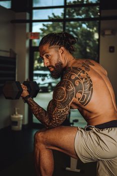 Tattooed man with beard working out indoors, lifting dumbbell, showcasing strength and fitness.