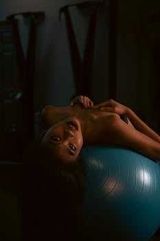 Elegant woman in a sensual pose on an exercise ball indoors, creating a moody and alluring atmosphere.