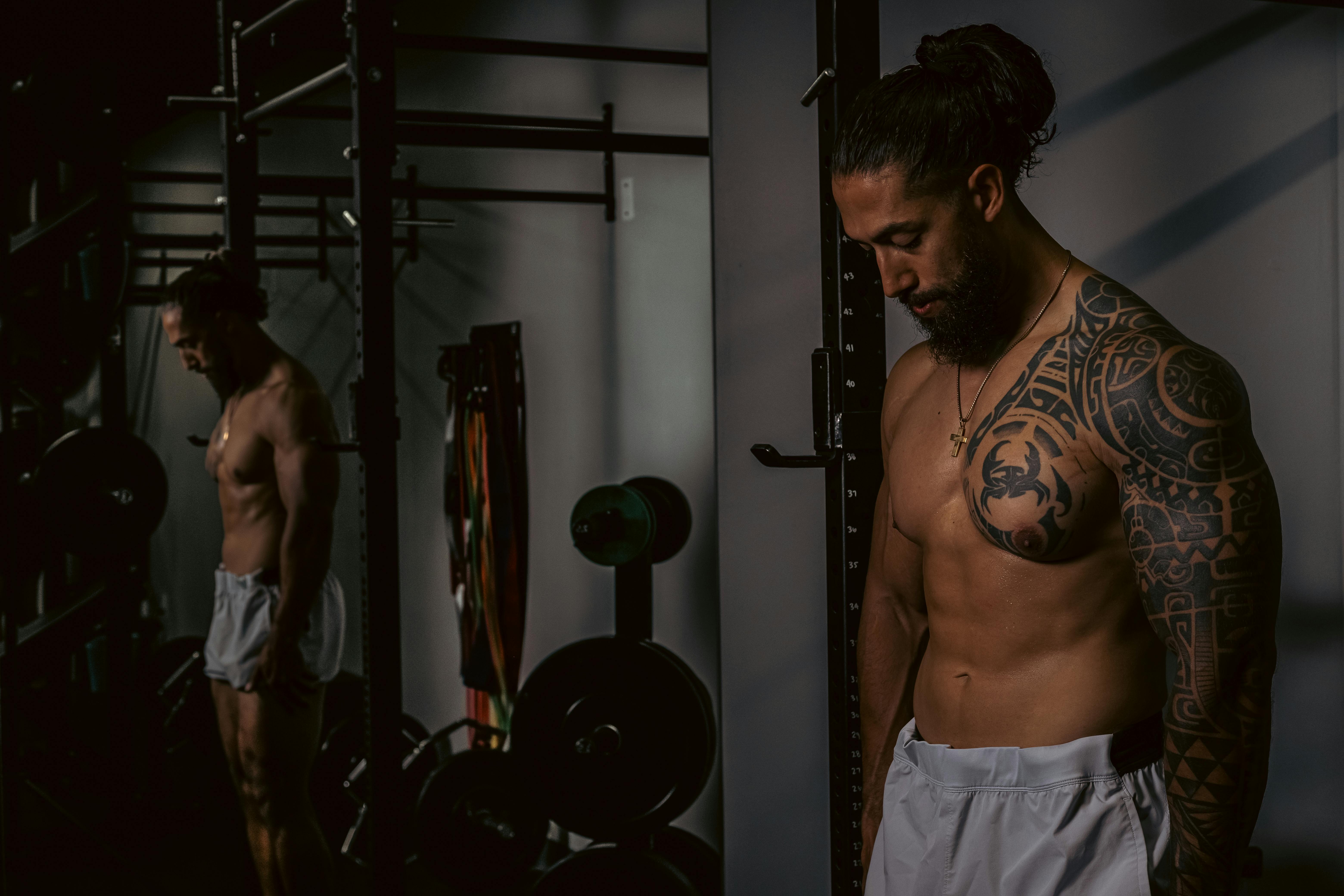 Shirtless muscular man with tattoos standing in a gym setting by the mirror.