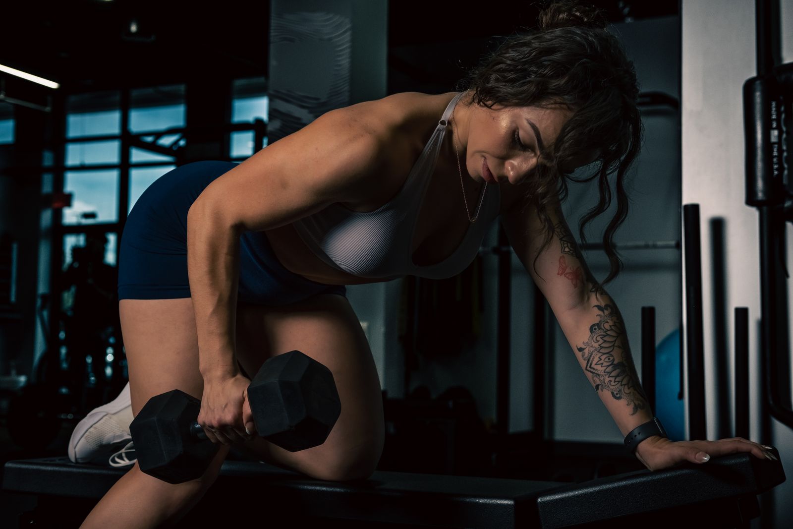 Woman with tattooed arm doing a one-arm dumbbell row on a bench in a dim gym