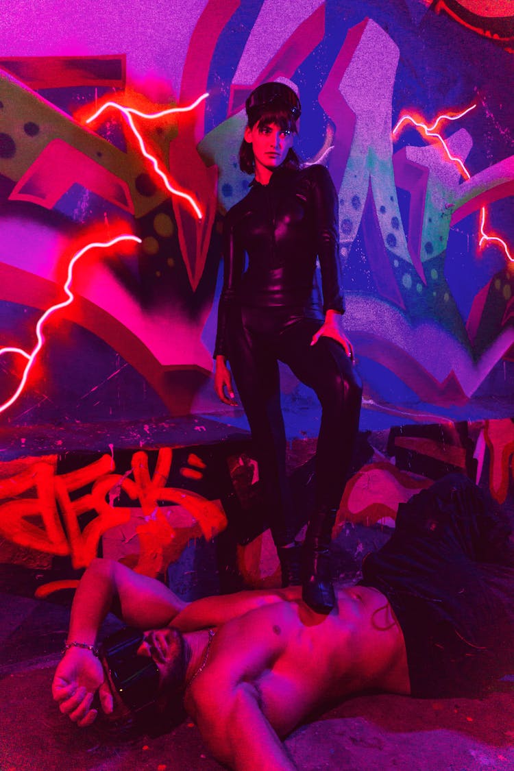 A Woman Wearing A Black Leather Suit Stepping On A Topless Man Near A Graffiti Background