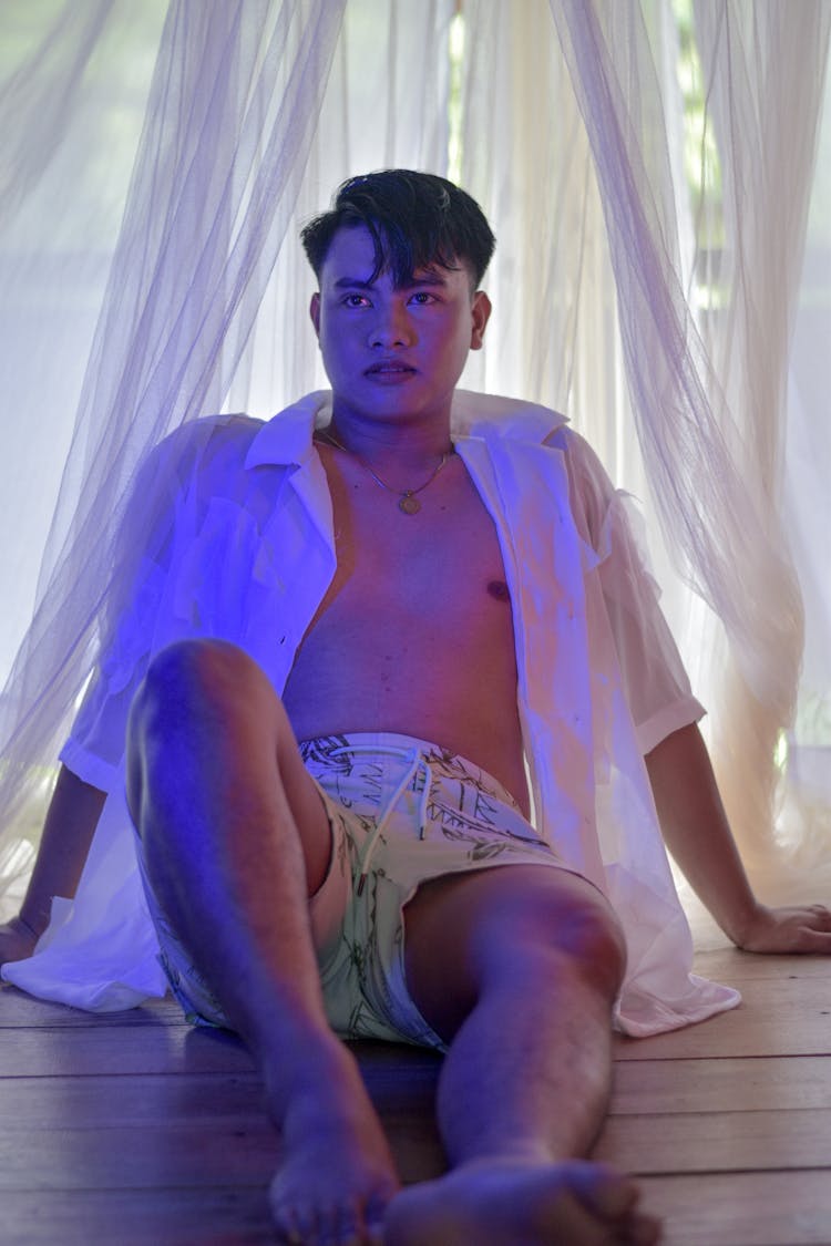 A Man Wearing Unbuttoned White Shirt And Boardshorts While Sitting On A Wooden Floor