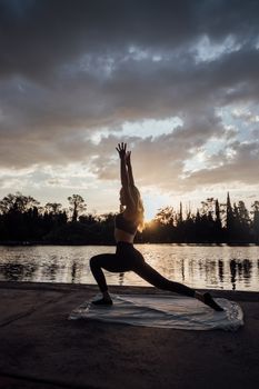 A woman performs yoga by a lakeside during a picturesque sunset, reflecting tranquility and fitness.