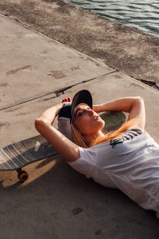 A young woman with blonde hair lies on concrete, holding a skateboard near a waterfront.