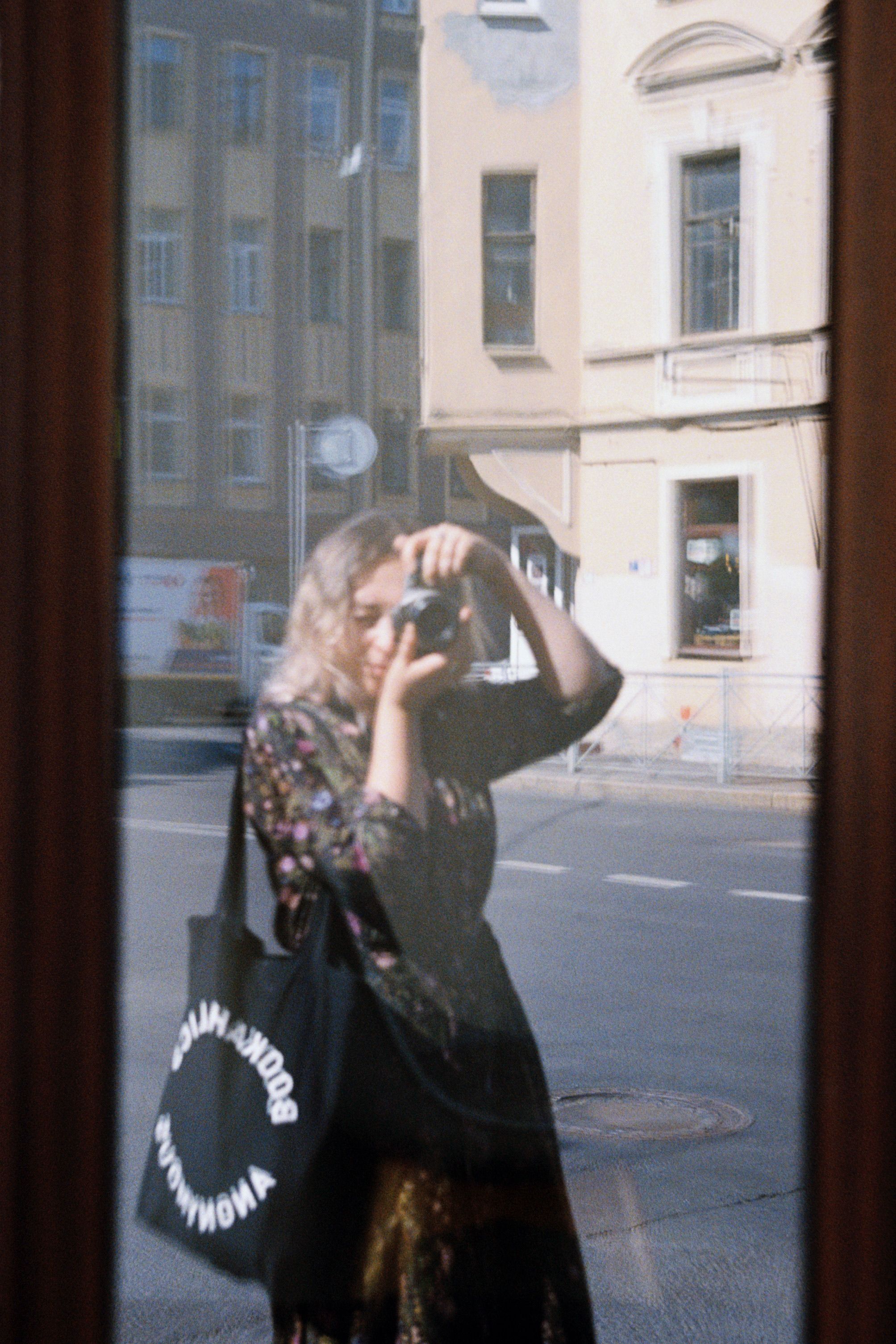A photographer takes a picture of their reflection in an urban window, capturing street life.